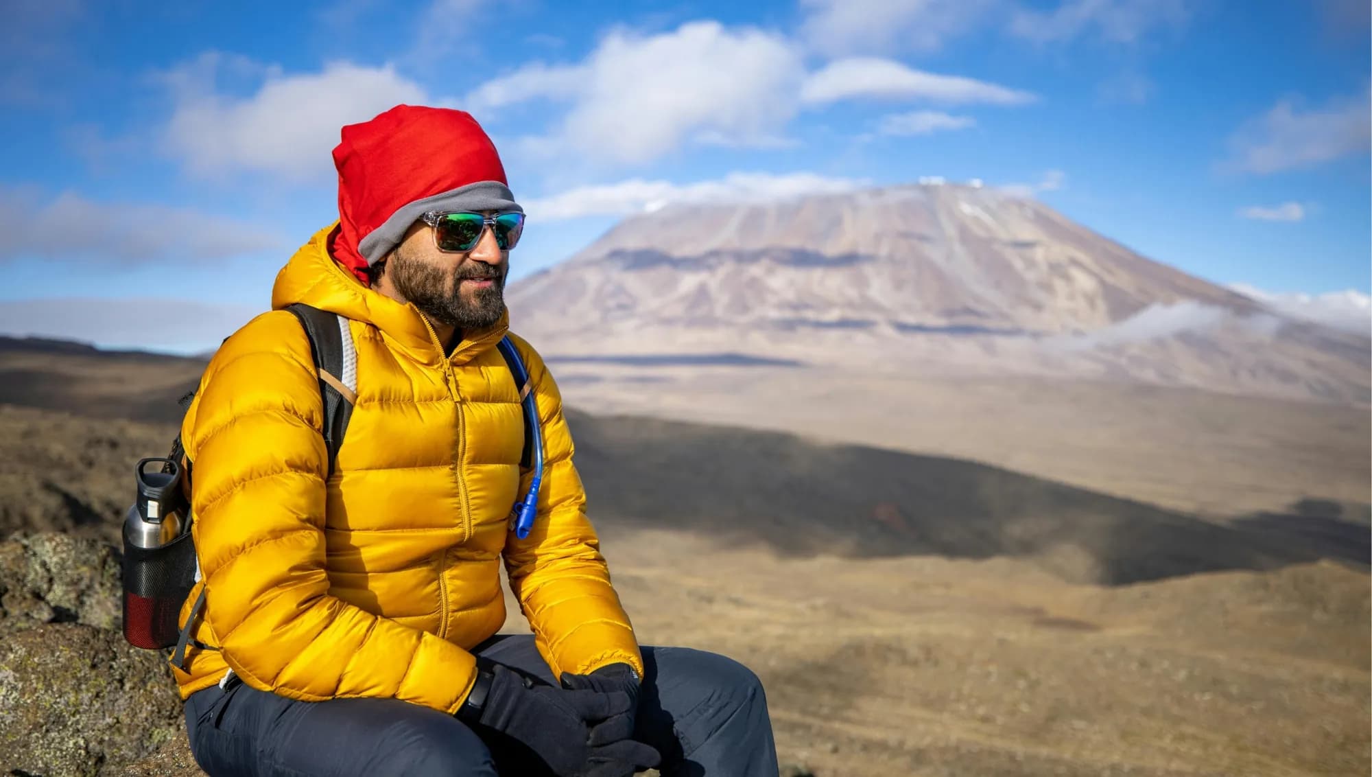 A climber in a down jacket resting on high ground with Kilimanjaro behind